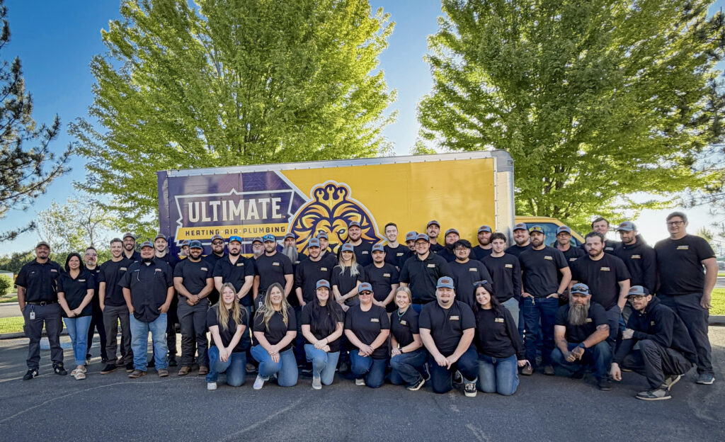A group of people in matching black shirts poses in front of a company truck with a yellow and purple Ultimate Heating & Plumbing logo. Trees are visible in the background.