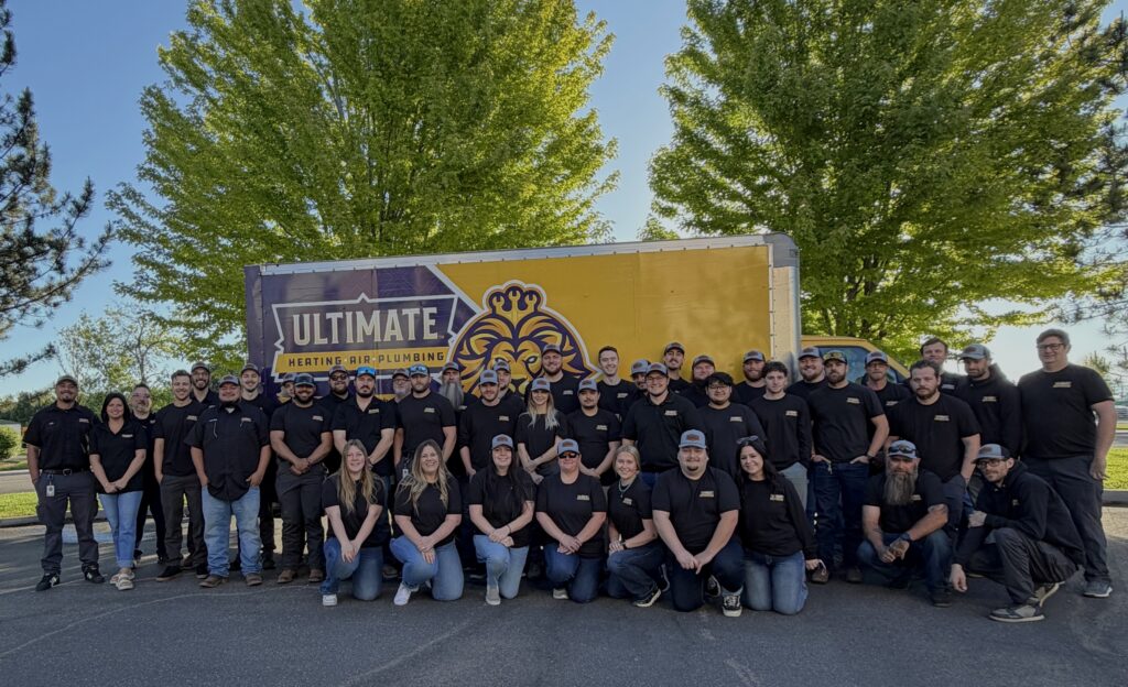 A large group of people wearing black shirts and caps pose in front of a truck with the "Ultimate Heating & Air Plumbing" logo, outdoors with green trees in the background.