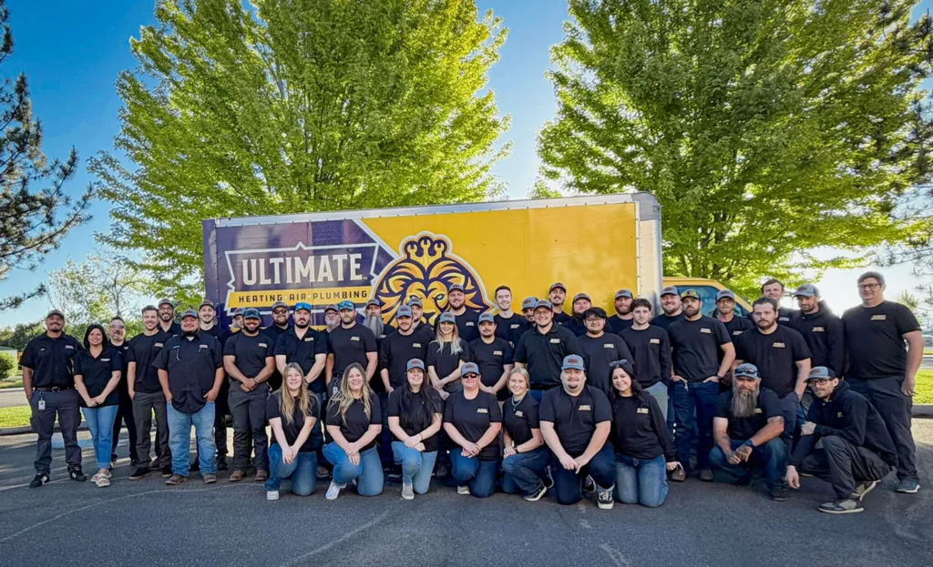 A large group of people in matching black shirts pose in front of a yellow Ultimate Heating, Air & Plumbing truck on a sunny day, with green trees in the background.